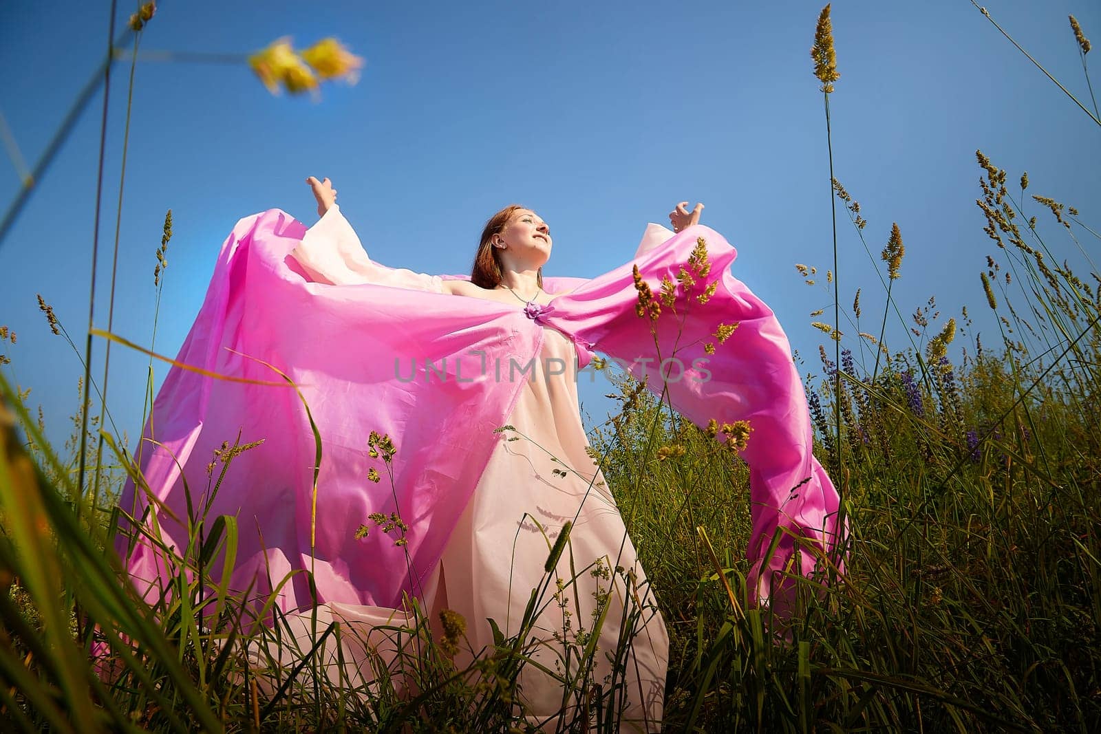 A woman joyfully spreads her arms in a vibrant pink dress, surrounded by lush greenery and blooms on a sunny day.