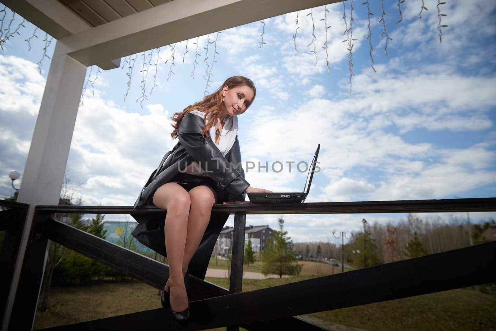 A woman sits confidently on a fence, using her laptop, with a picturesque landscape and cloudy sky in the background.