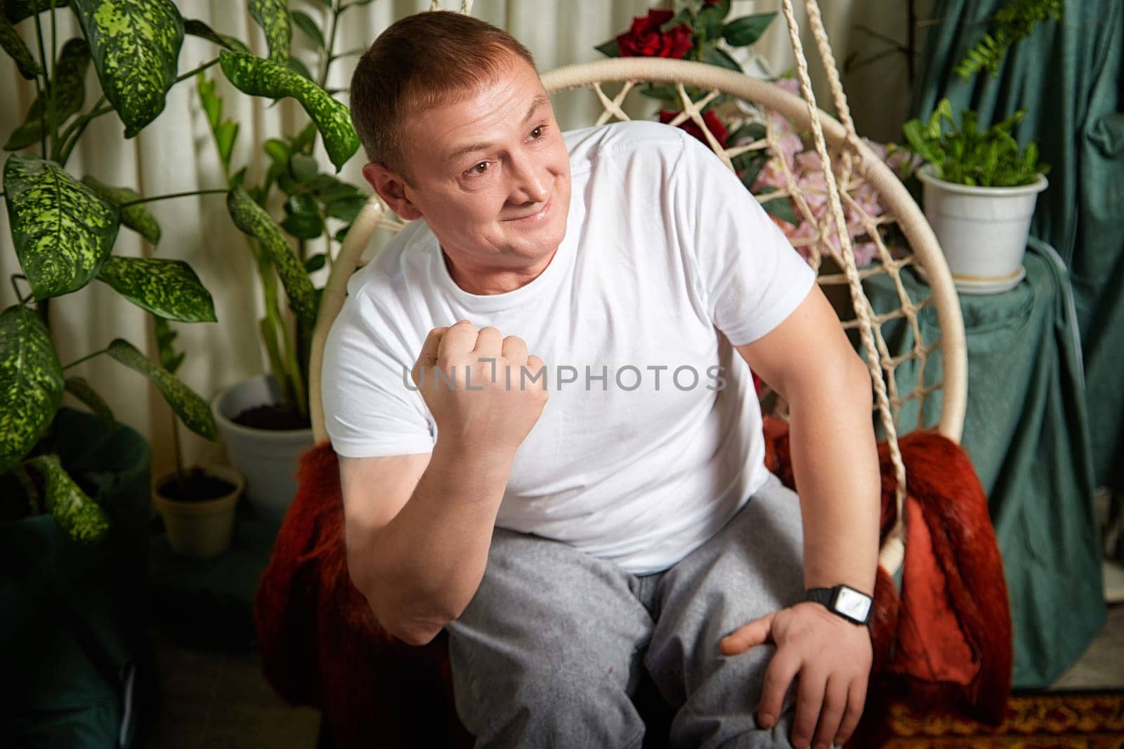 A man is sitting on a swing, smiling and showing enthusiasm while enjoying a relaxed moment in a plant-filled room.