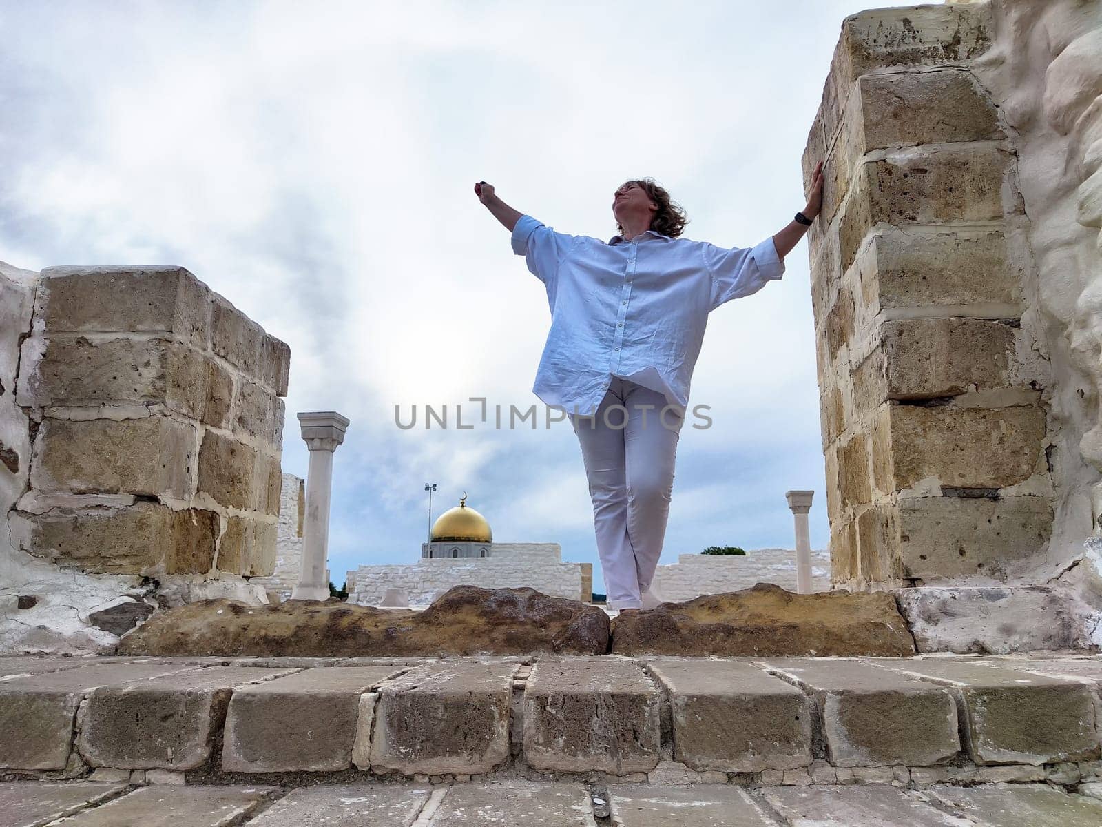 Blonde woman tourist stands confidently amidst ancient ruins, arms outstretched, embracing the adventure of exploring a historic location.