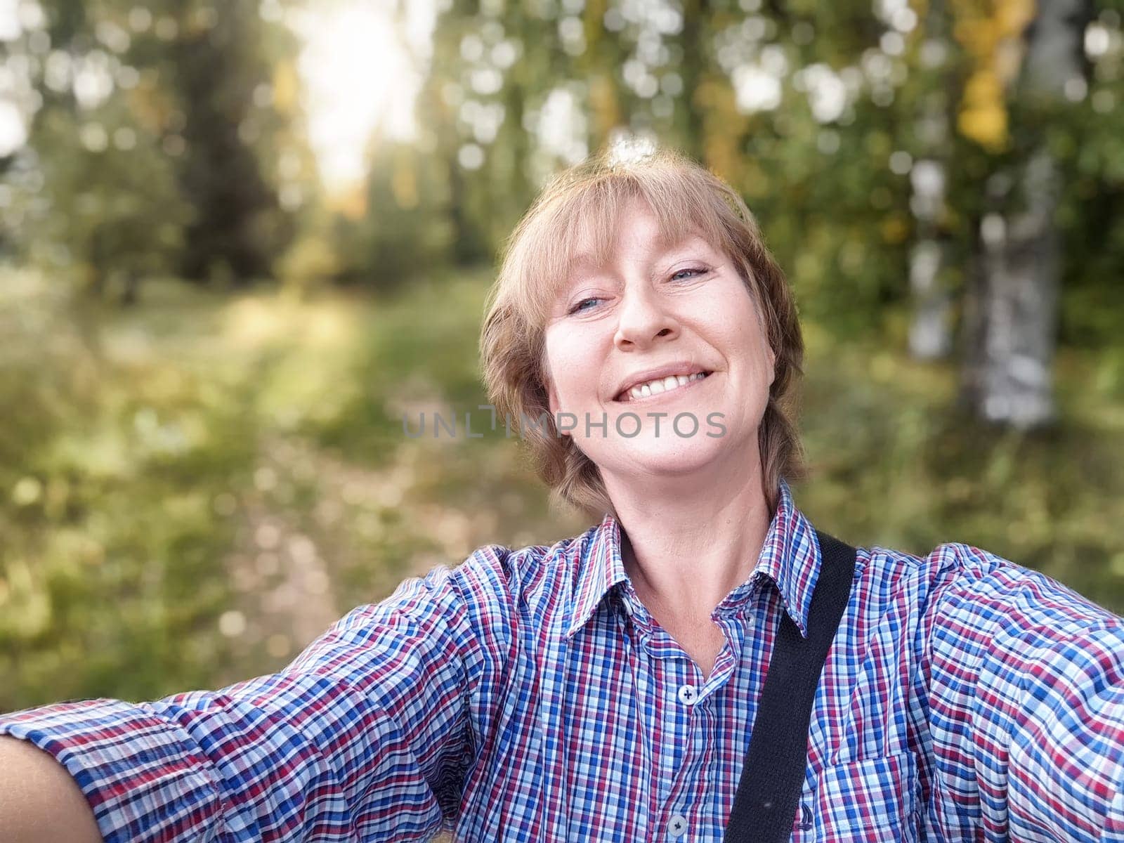 A middle-aged woman traveler captures a joyful selfie while exploring nature on a beautiful day in the forest.