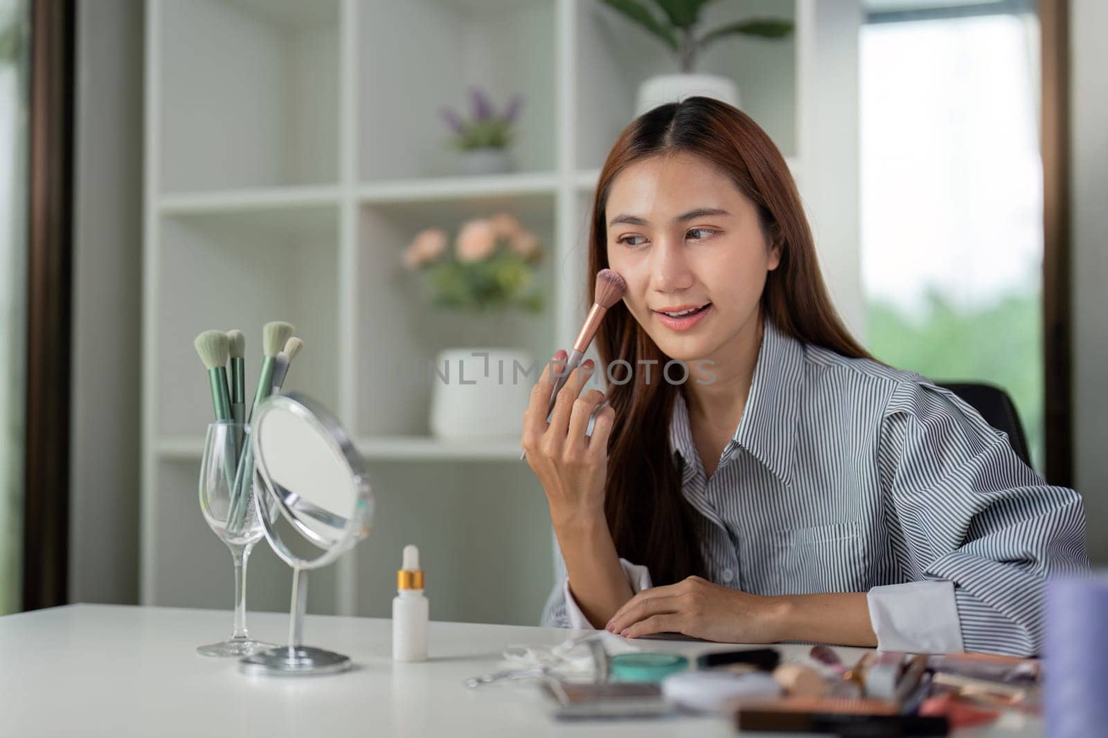 A woman focuses on her makeup application, skillfully applying blush with brush in front of a mirror. The serene environment enhances her beauty routine.