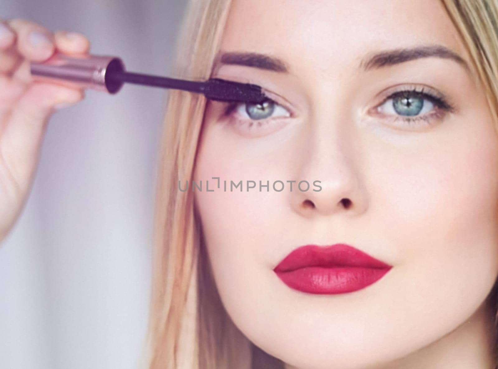 Woman applying mascara with confidence, defined brows and red lipstick. Blonde beauty focused on her eye makeup, cosmetics look