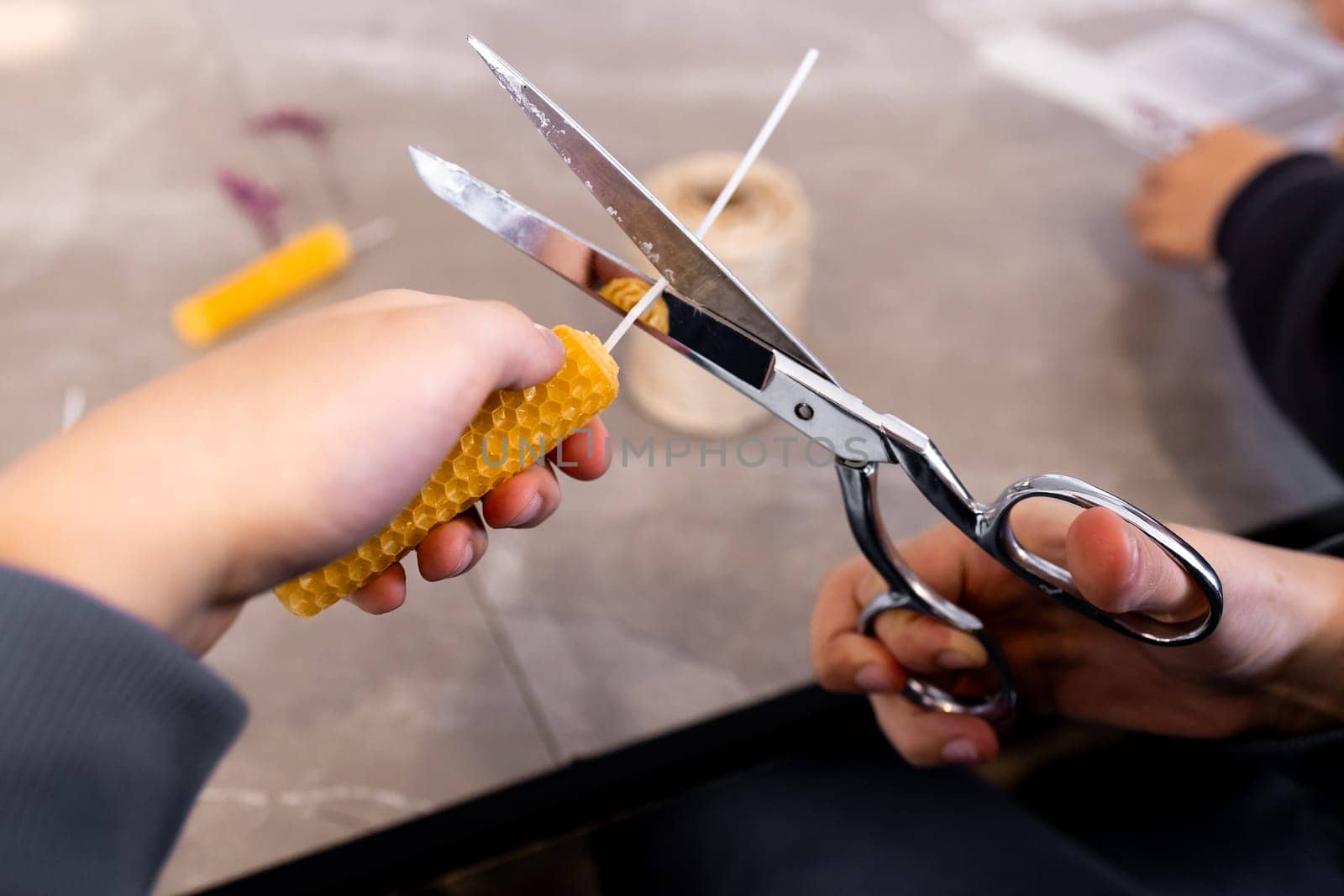 Hands rolling beeswax candle with scissors and wick on table.