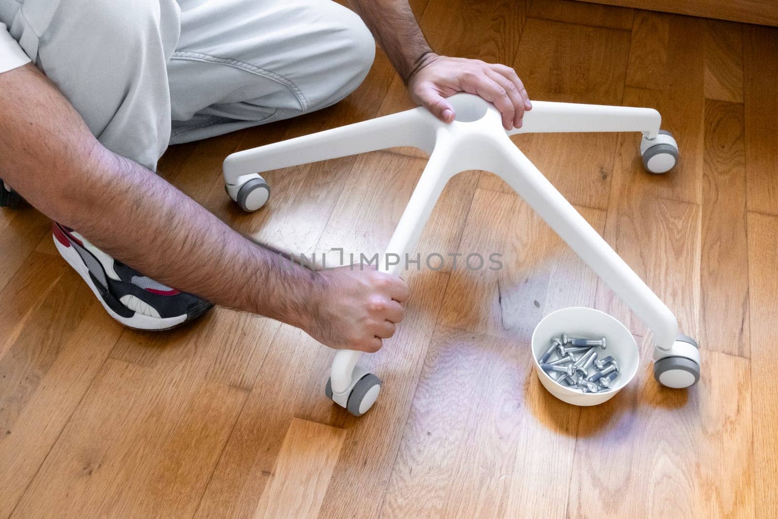 Male adult assembling office chair base with wheels on wooden floor.
