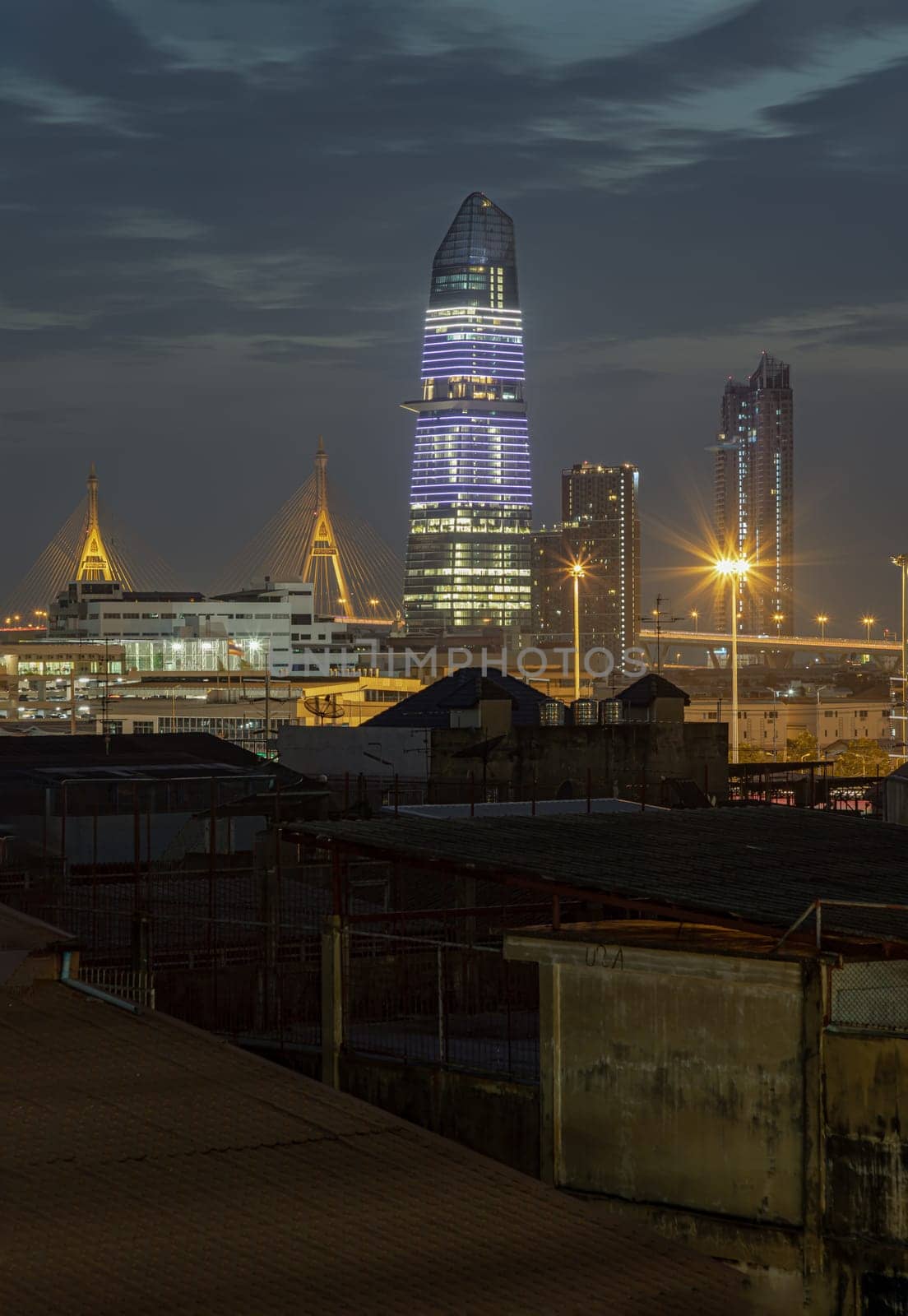 Bangkok, Thailand - Dec 04, 2024 - Stunning view of KingBridge tower alongside the iconic landmark of Bhumibol Bridge and Skyscrapers in the business district of Bangkok city with bright glowing lights in the night. Architectural of Modern buildings, use it as your Wallpaper, Poster and Space for text, Selective focus.