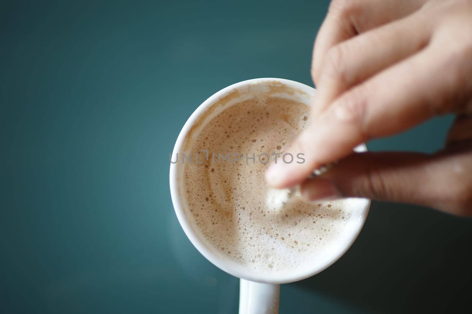 A hand stirs a foamy drink in a white mug on a green surface.
