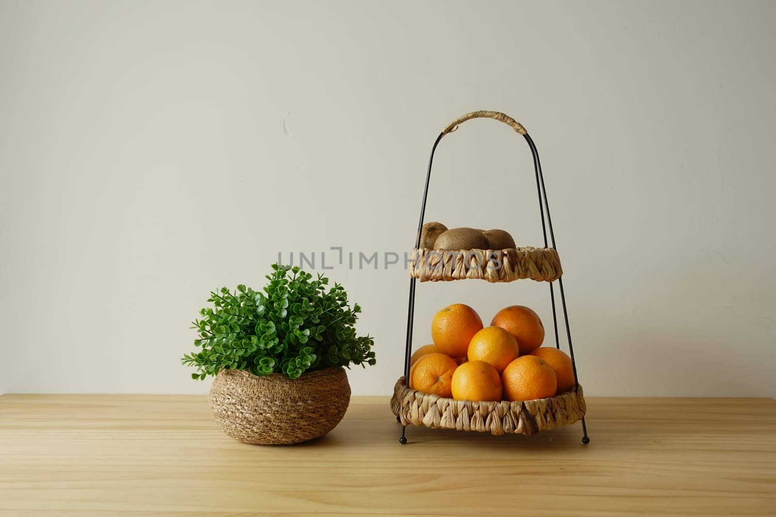 A two-tiered basket holds vibrant oranges and kiwis beside a potted plant.