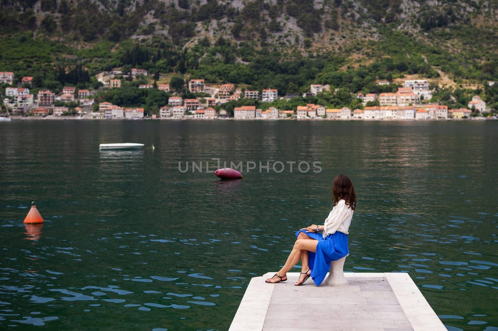 A woman in a blue skirt sits on a pier, overlooking a picturesque bay with houses on the hillside.