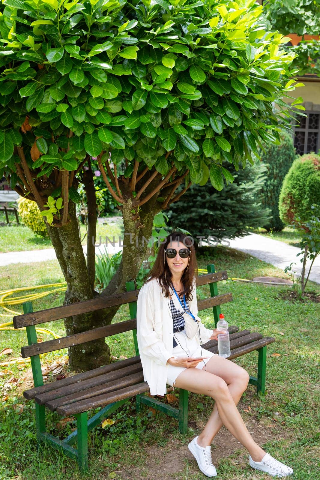 A young woman relaxes on a park bench under a lush green tree, enjoying a drink and her music.