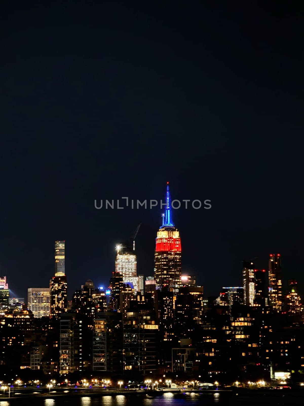 The Empire State Building glows in red, blue, and gold above the New York City skyline, reflecting in the calm nighttime river.