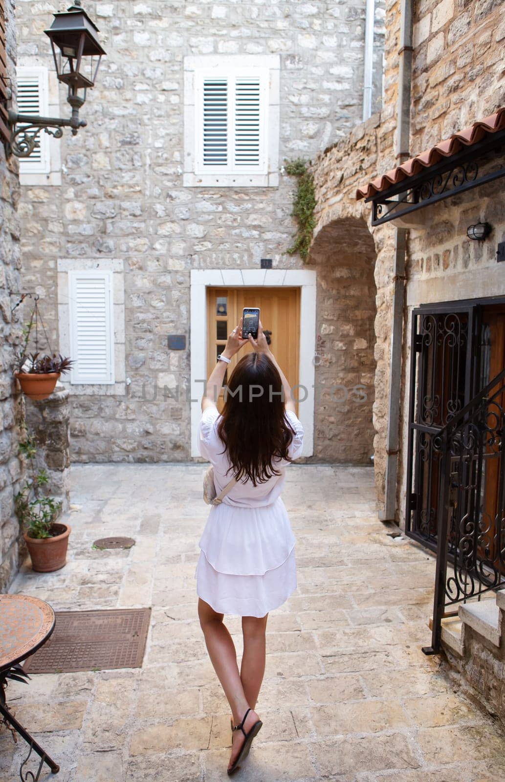 A woman in a white dress takes a photo on her phone in a quaint stone alley. Travel in Europe