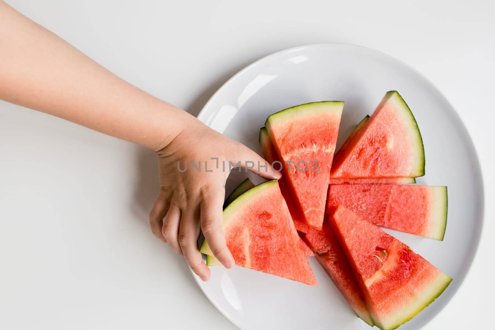 Child hand reaching for sliced watermelon on white plate.