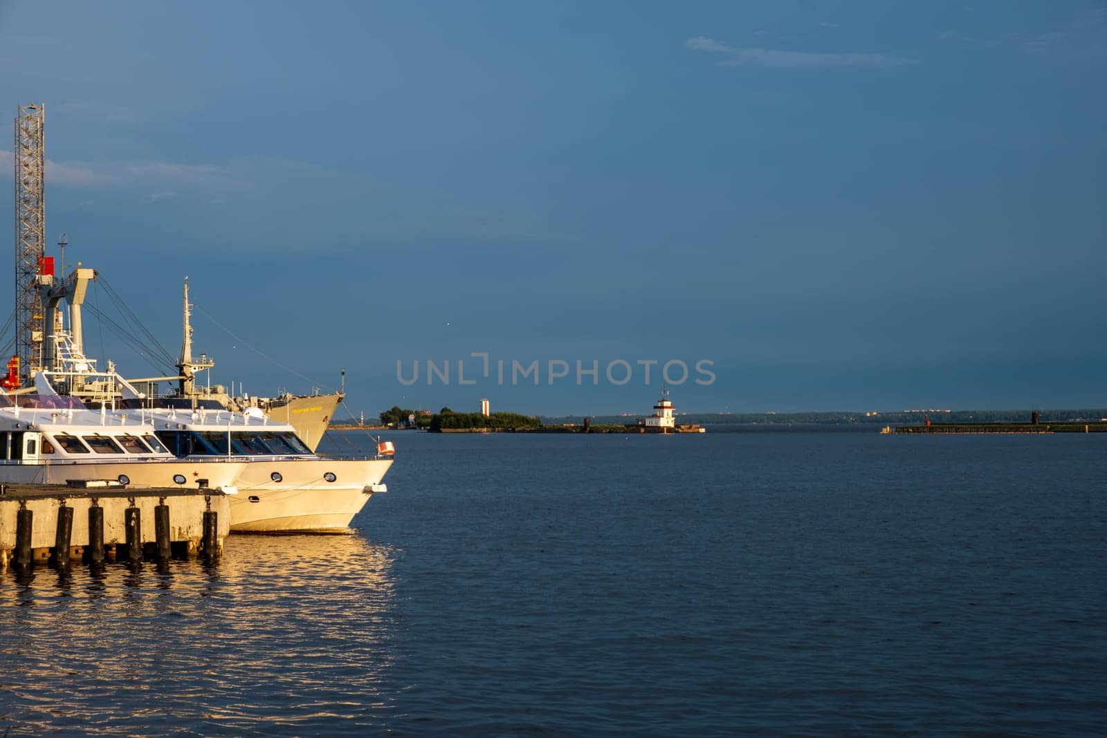 Kronstadt, Russia - 07.27.24. Serene harbor view featuring boats docked at a pier, with a lighthouse visible in the distance. Tranquil setting of early evening.