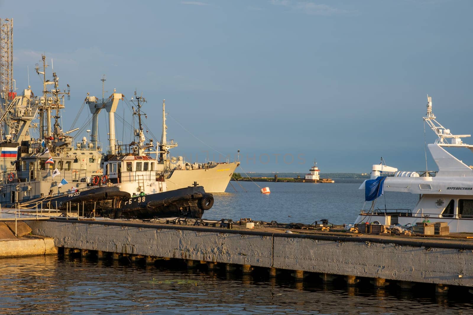 Kronstadt, Russia - 07.27.24. Serene harbor scene featuring various ships docked at the pier, with a lighthouse visible in the distance. The golden hour lighting.