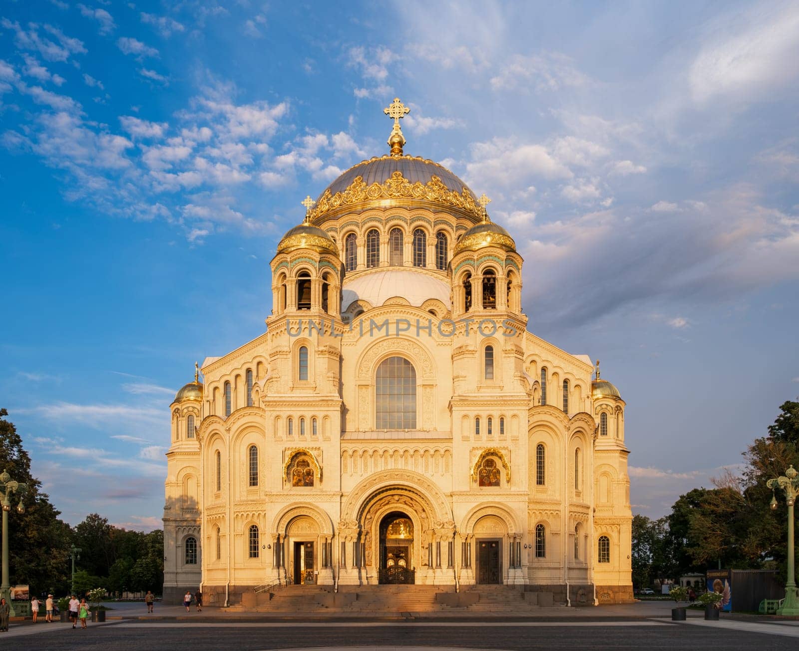 Kronstadt, Russia - 07.27.24. Naval cathedral facade illuminated by evening sunlight, showcasing intricate architectural details and golden domes against a blue sky.