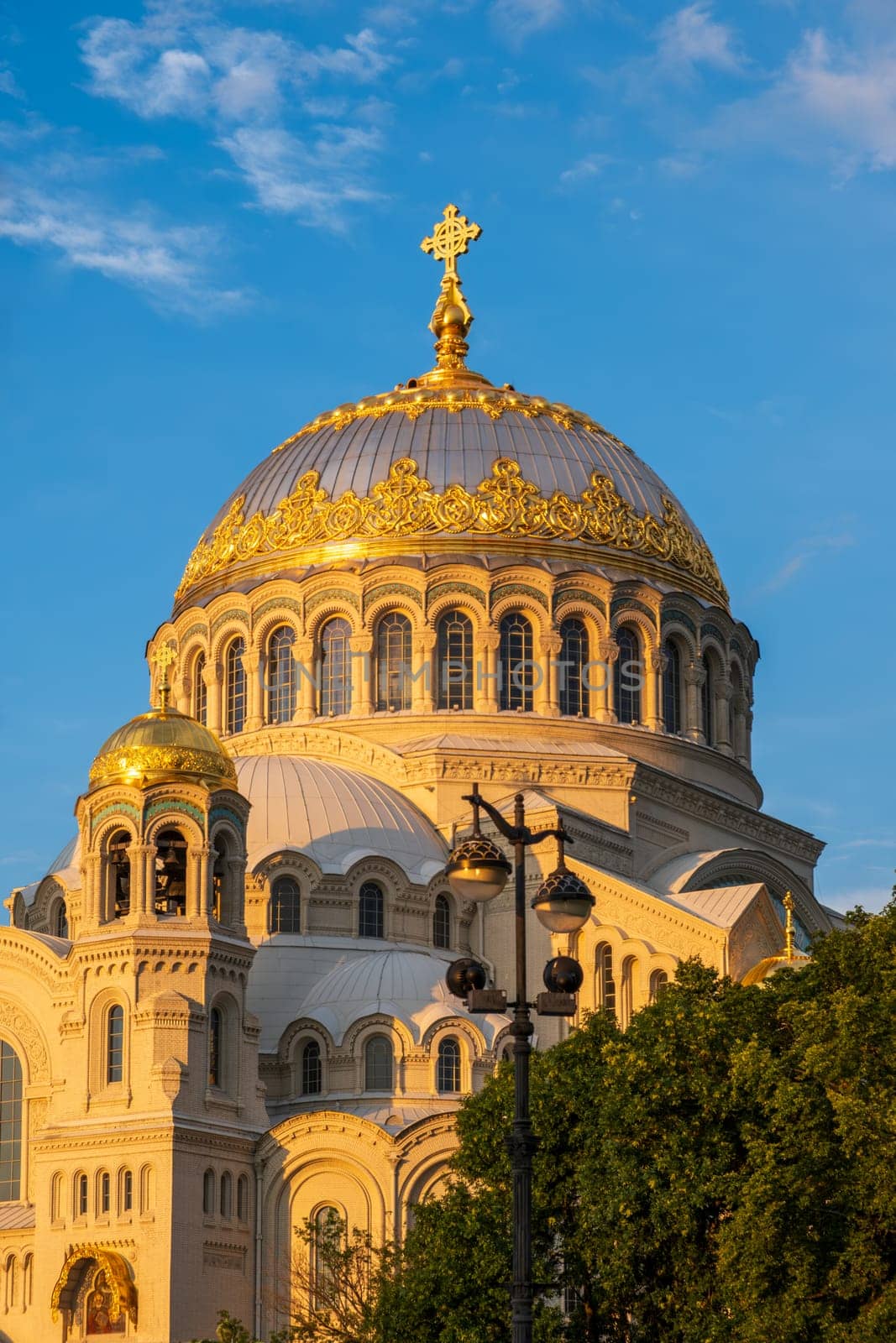 Kronstadt, Russia - 07.27.24. Naval cathedral facade illuminated by evening sunlight, showcasing intricate architectural details and golden domes against a blue sky.
