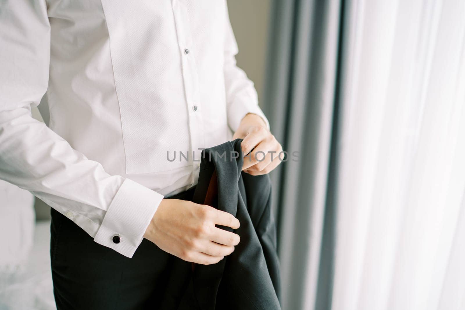 Groom holds a black jacket in his hands while standing in a hotel room. Cropped. Faceless. High quality photo