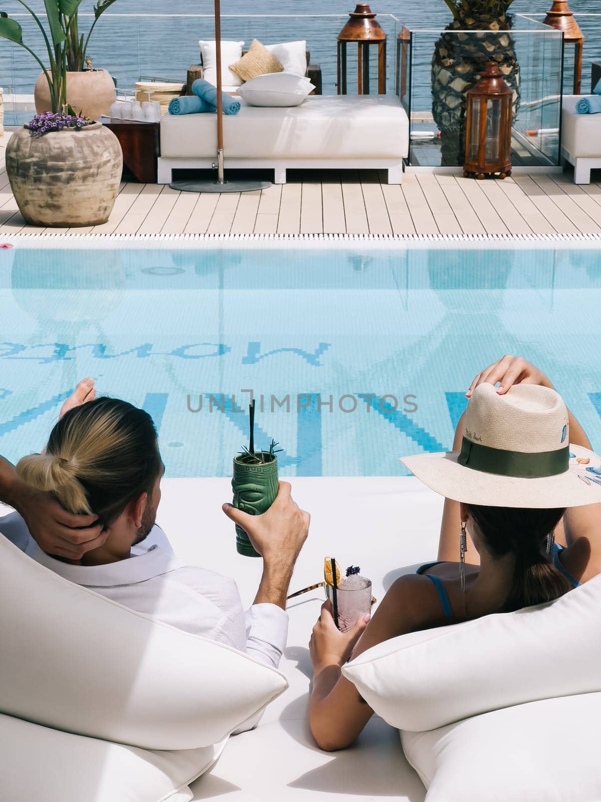 Man and woman with cocktails lie on a double deck chair by the pool. Back view. High quality photo