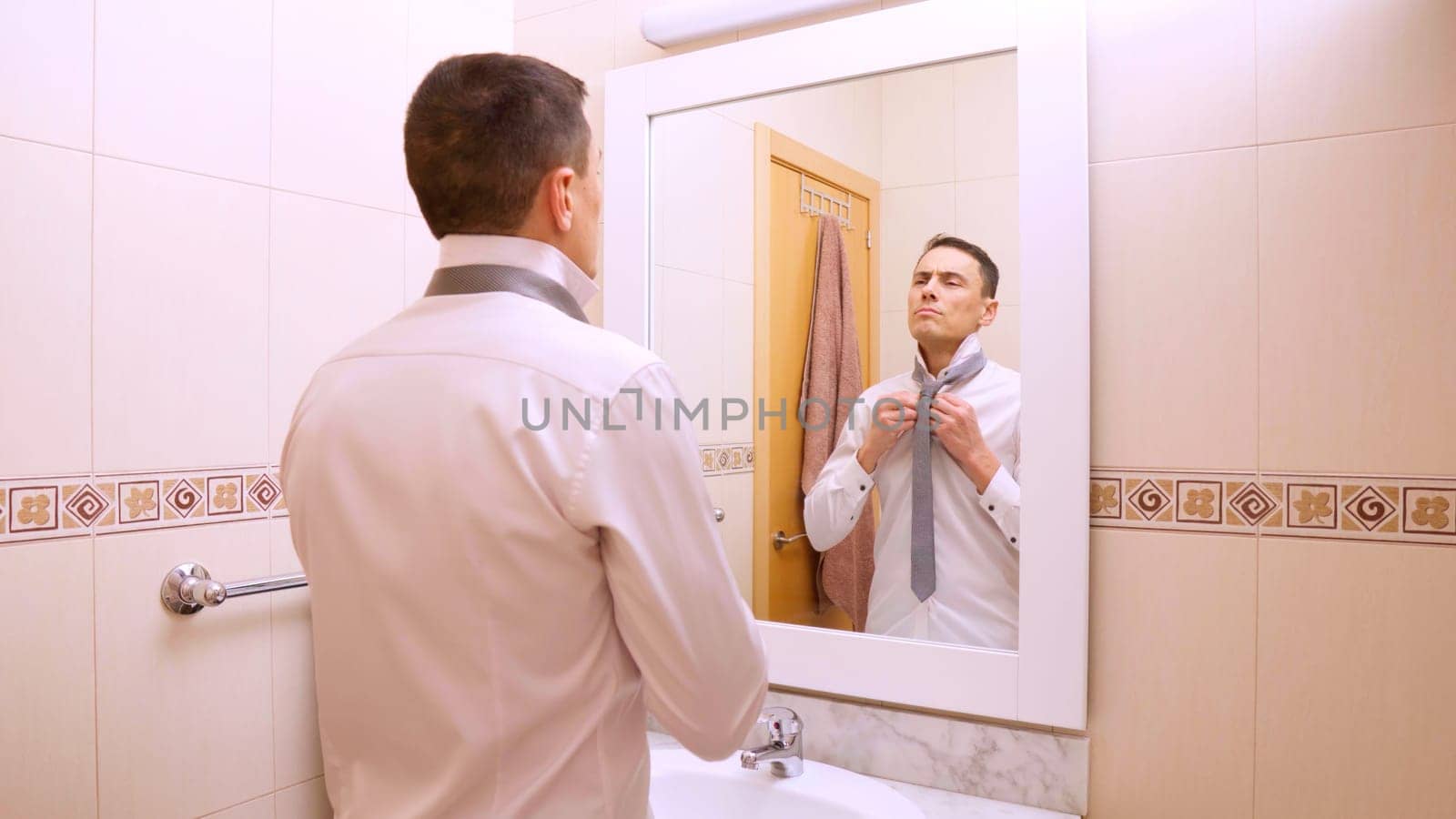 Businessman is getting ready in the morning, buttoning his shirt and adjusting his tie in front of the bathroom mirror