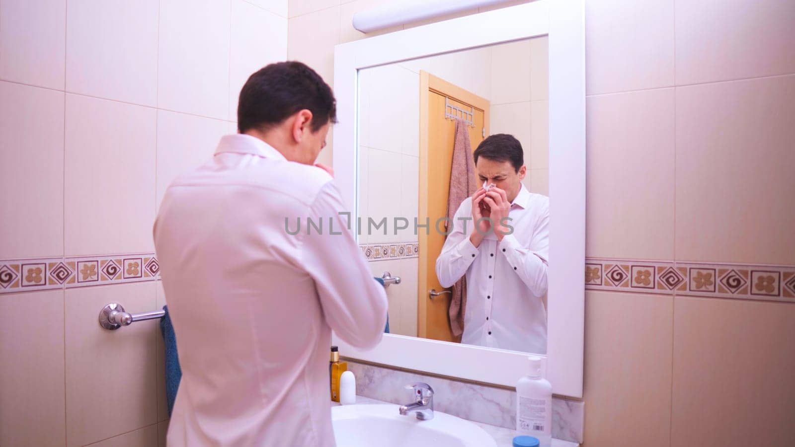 Office worker blowing his nose with a tissue while looking in the bathroom mirror