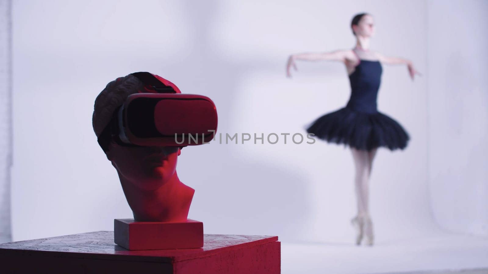Young woman ballerina in black dress training - a head of greek sculpture in VR glasses on the foreground - red lighting. Mid shot