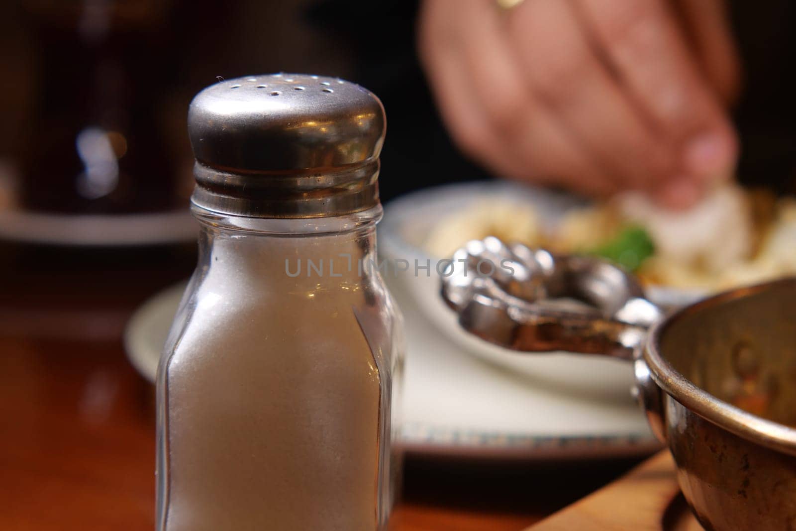 A salt shaker rests on a dining table as a hand reaches for food.