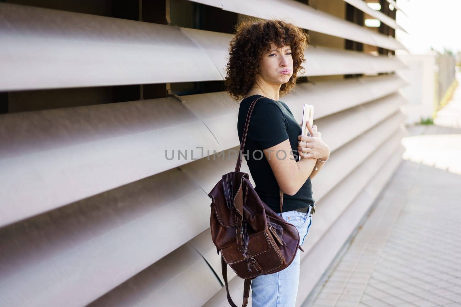 A young woman with beautiful, curly hair is confidently standing against a sleek, modern wall. She holds a phone in one hand and a stylish bag in the other, showcasing a trendy and expressive look