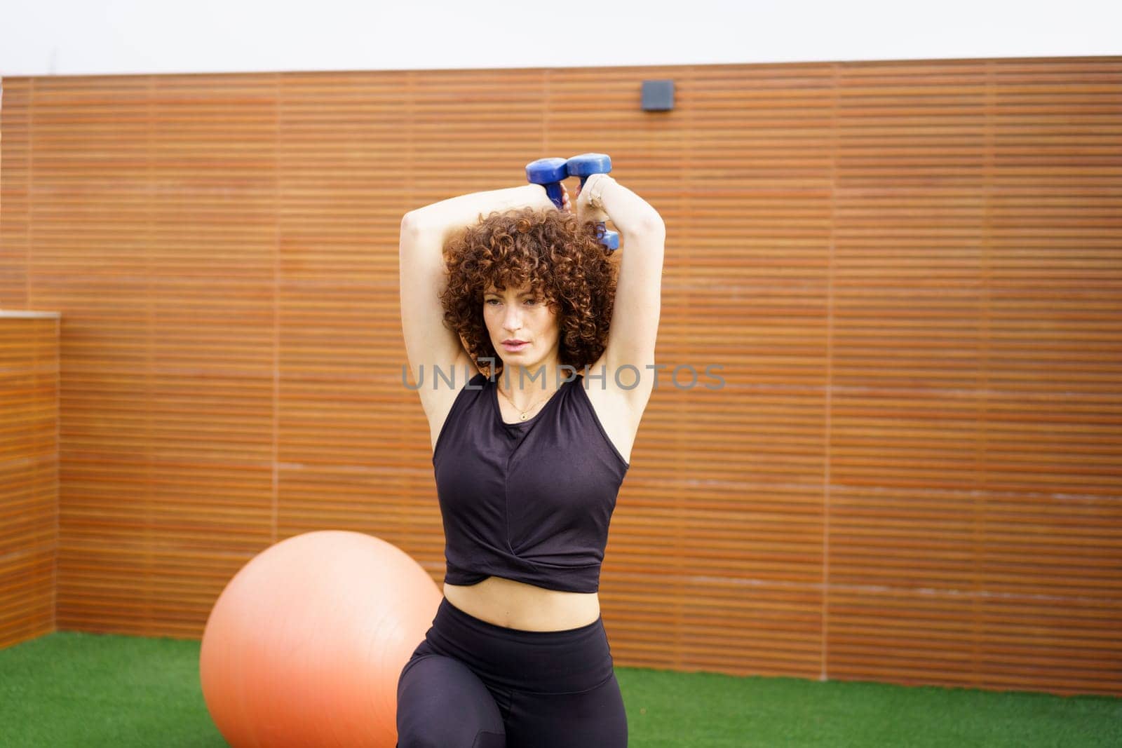 A woman with beautiful, curly hair is actively performing strength training exercises with dumbbells in a stylish and modern gym environment, which features an impressive large exercise ball