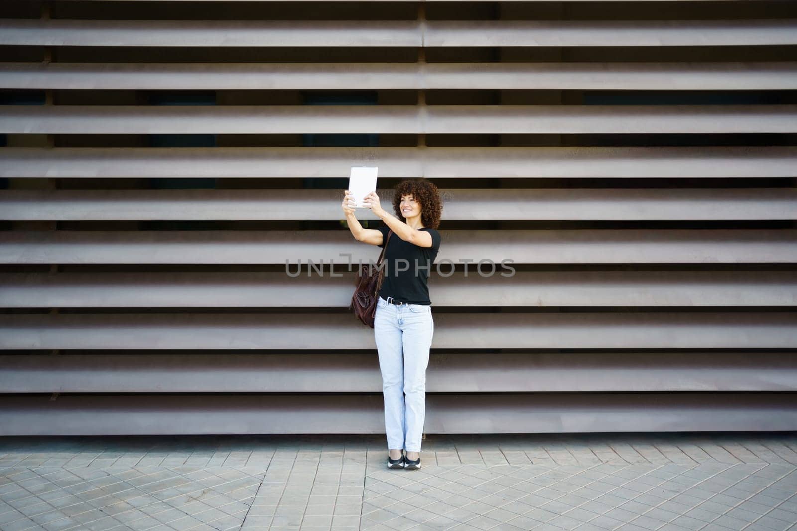A joyful woman with curly hair holds her smartphone high, ready for a selfie in front of an elegant wall, capturing the vibrant essence of urban life and the pure joy from her radiant smile