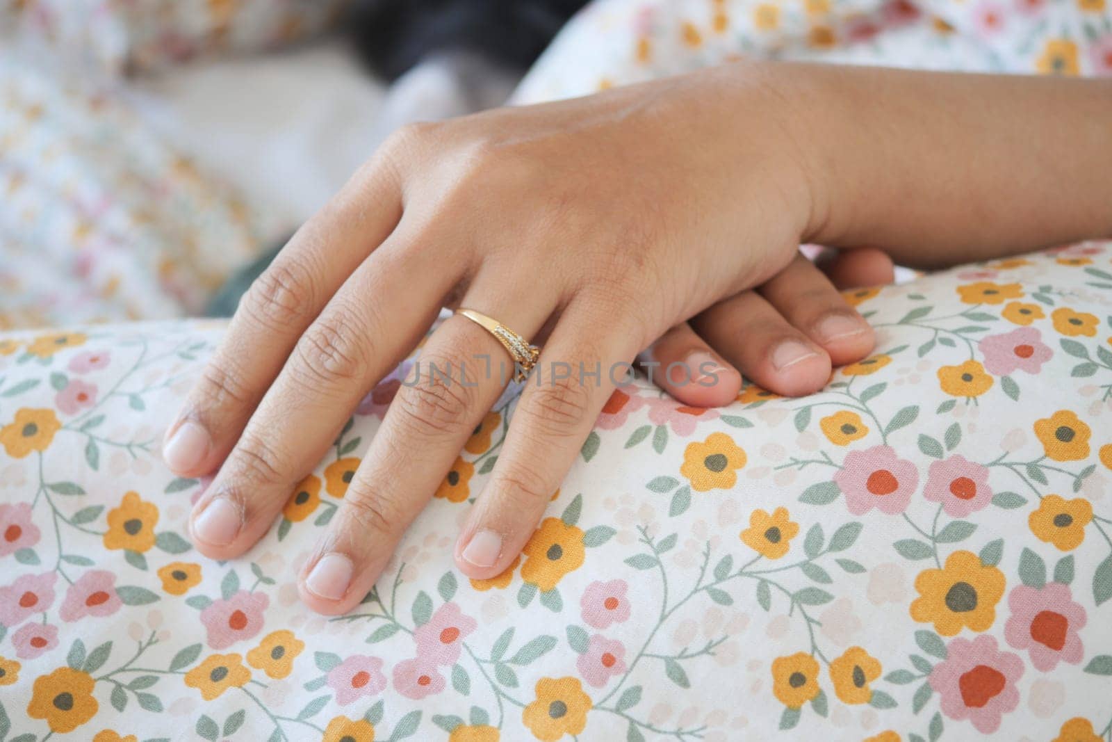 A hand adorned with a ring rests gently on patterned bedding in sunlight.