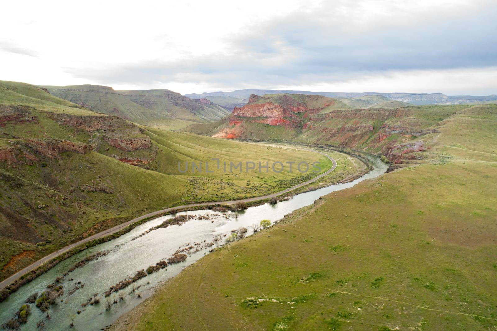 A river runs through a grassy plain with a rocky mountain in the background. The sky is cloudy and the landscape is serene