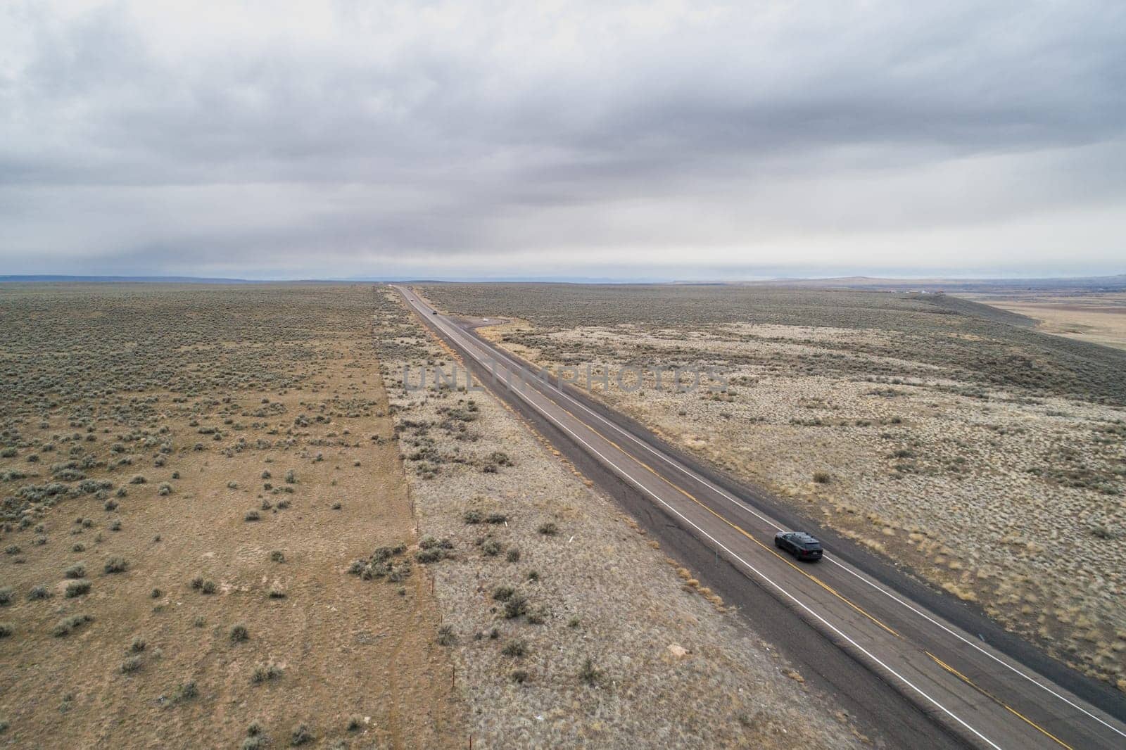 A car is driving down a long, empty road in a desert. The sky is cloudy and the landscape is barren