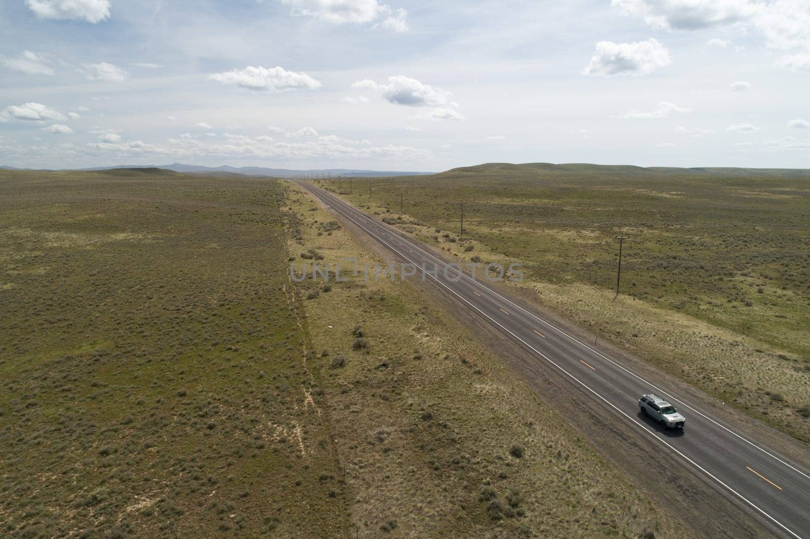A car is driving down a long, empty road in a desert. The sky is cloudy and the landscape is barren