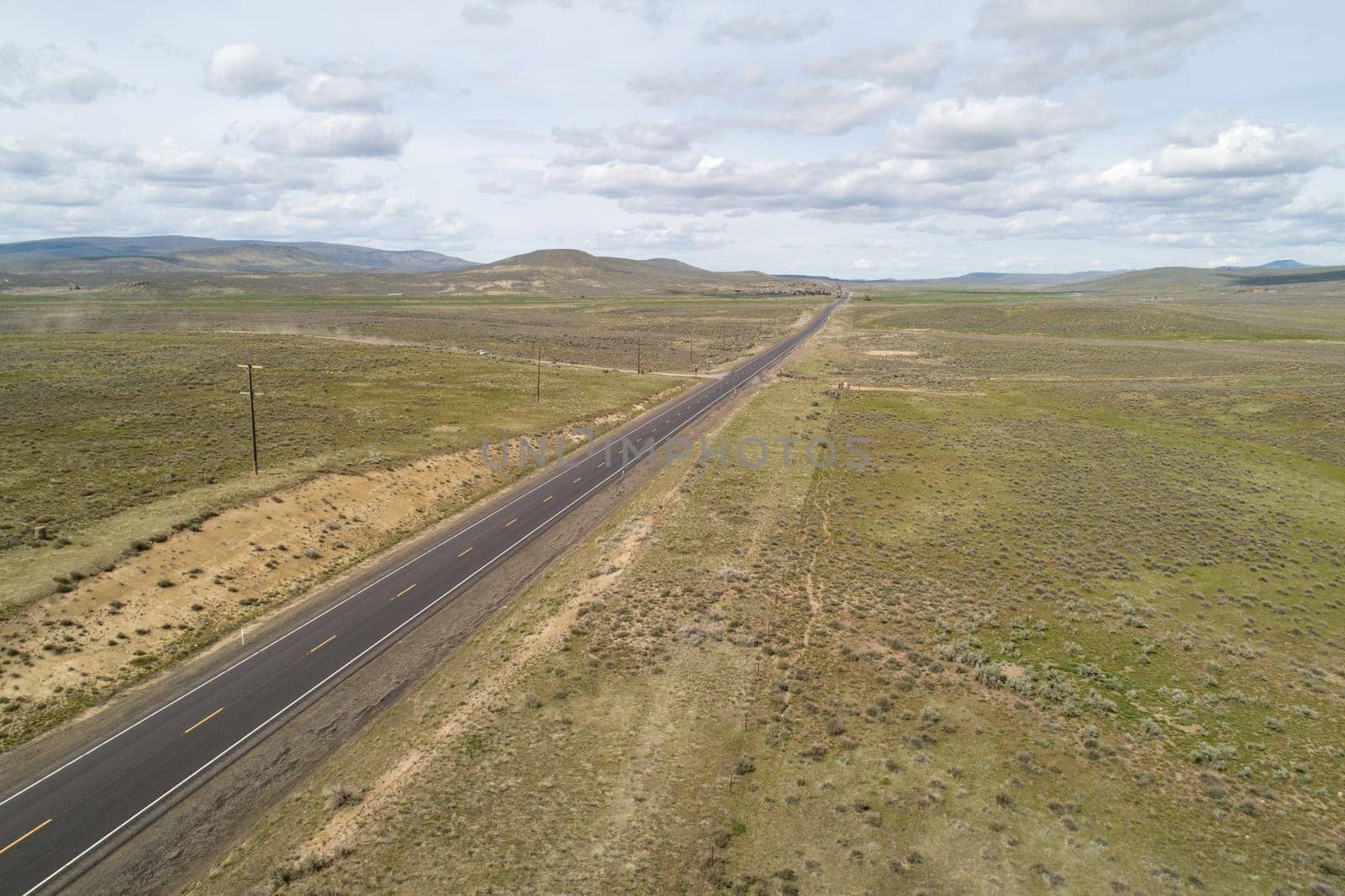 A long, empty road stretches across a dry, barren landscape. The sky is cloudy, and the sun is barely visible. The scene is desolate and quiet, with no signs of life or activity