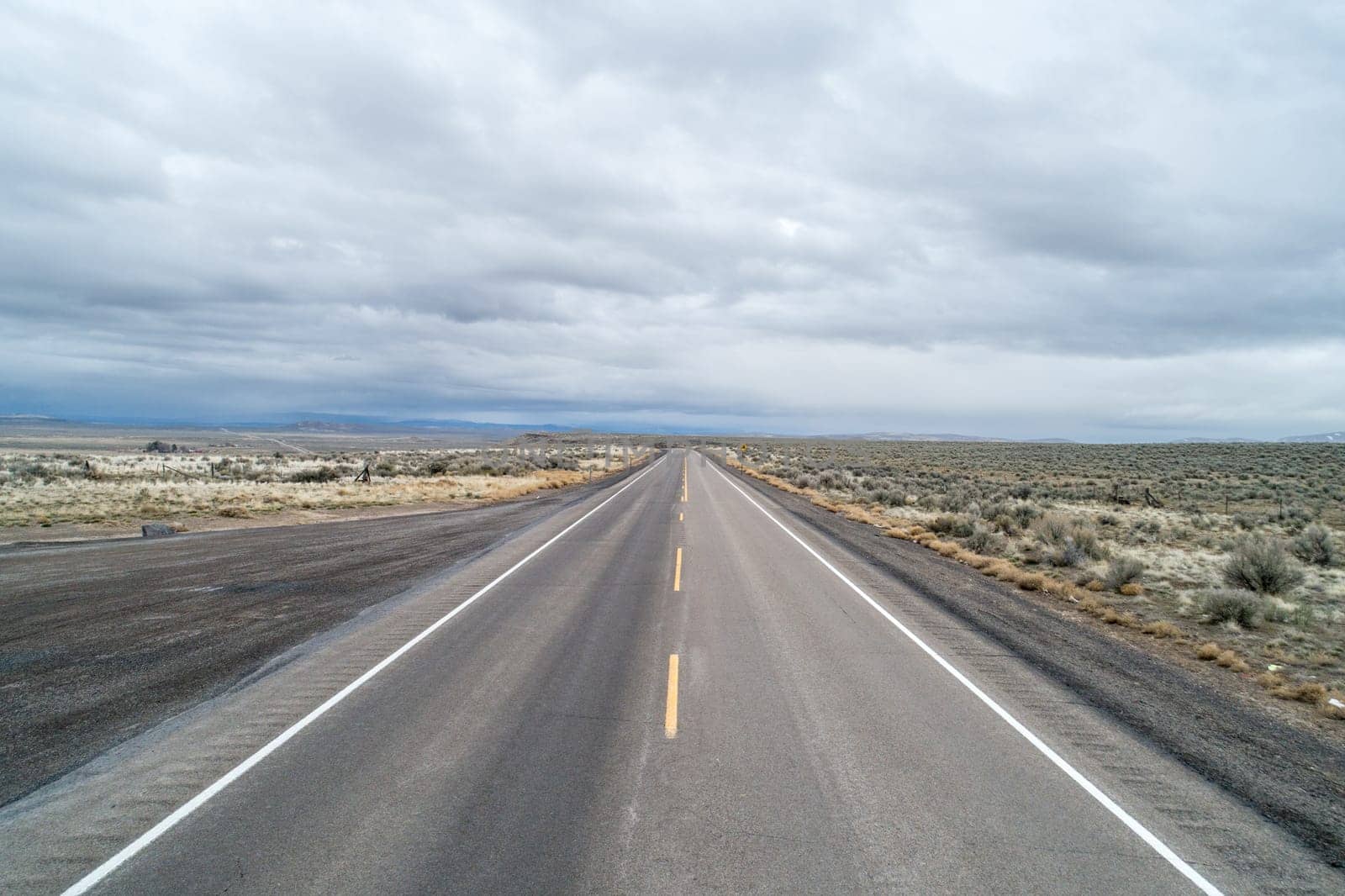 A long, empty road with a few trees in the distance. The sky is cloudy and the road is wet