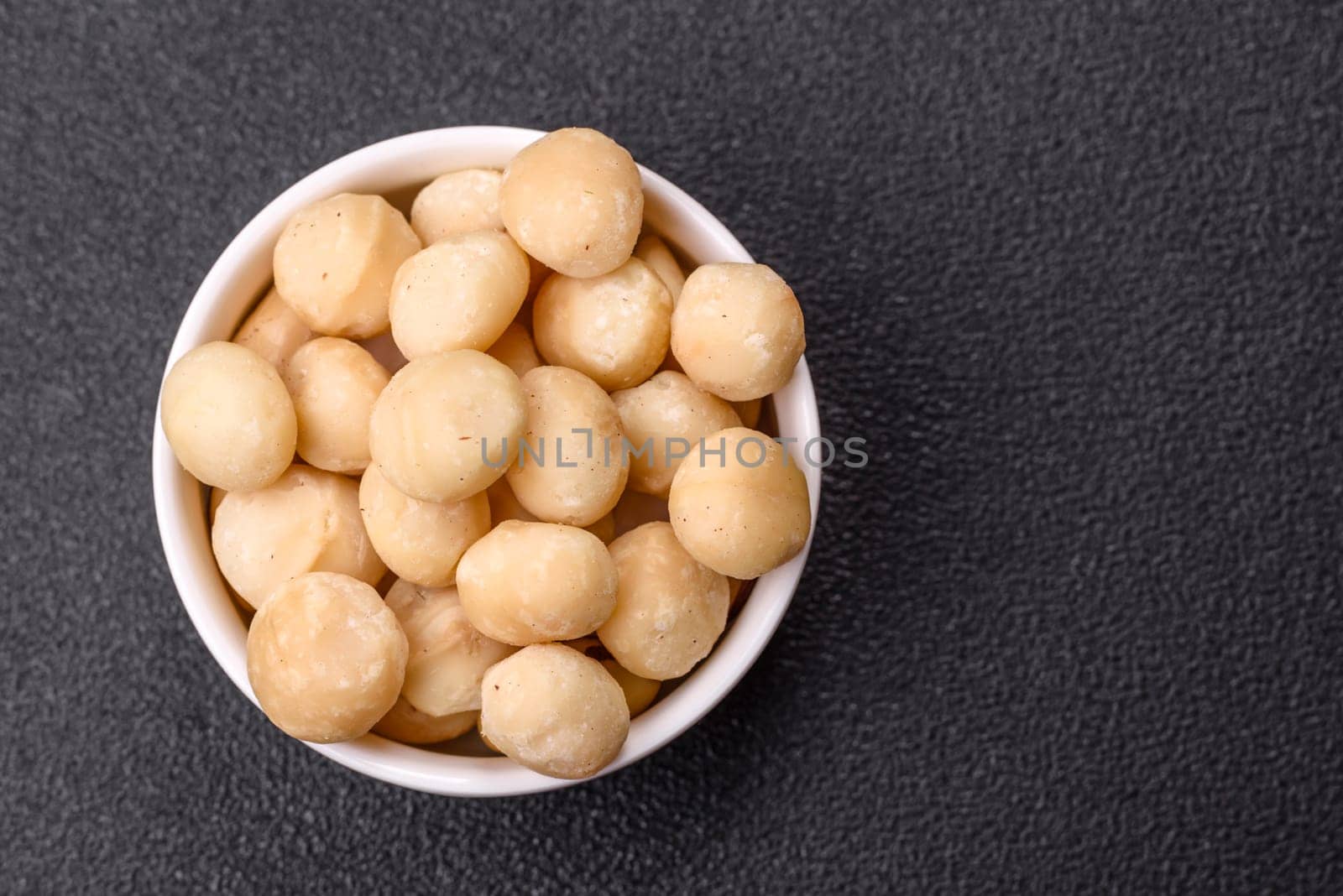 Raw peeled macadamia nut in ceramic bowl on dark concrete background. Nuts as a source of nutrients