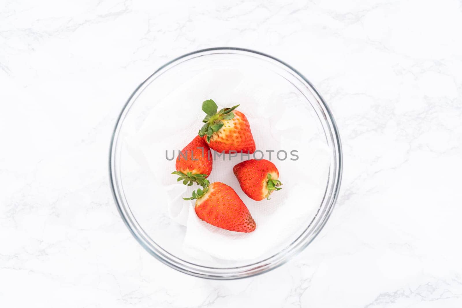 Bright red strawberries, interspersed with signs of mold, rest in a glass bowl lined with a paper towel on a white napkin, indicating improper storage techniques.