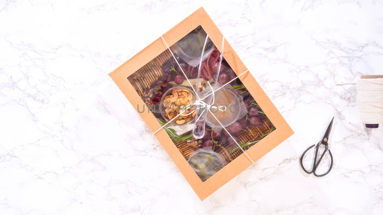 Flat lay. Hands are meticulously adding fresh red grapes to a bowl, complementing a beautifully arranged charcuterie box brimming with a variety of cheeses, olives, and cured meats, set against a sleek marble surface.