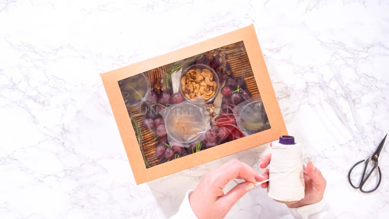 Flat lay. Hands are meticulously adding fresh red grapes to a bowl, complementing a beautifully arranged charcuterie box brimming with a variety of cheeses, olives, and cured meats, set against a sleek marble surface.