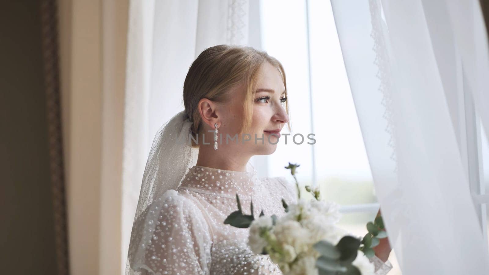 Beautiful bride in white wedding dress holding bouquet and looking out window on her wedding day