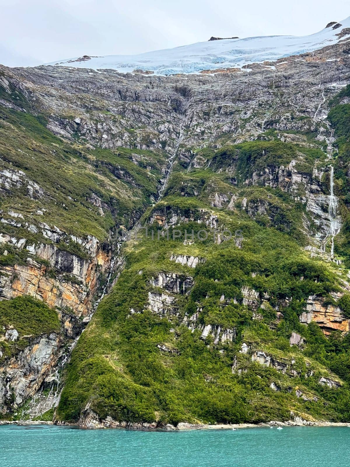 Thin glacial waterfalls stream down rugged, forested cliffs from an icy glacier above, reaching the vivid turquoise waters of a remote fjord below.