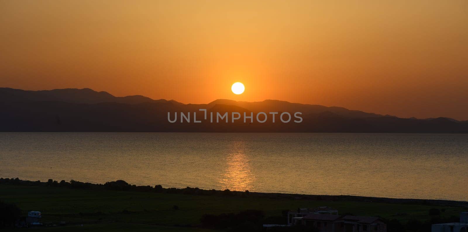 As the sun descends, the sky transforms into warm shades of orange and gold, casting a peaceful glow over calm waters and silhouetted mountains, creating a breathtaking evening view.