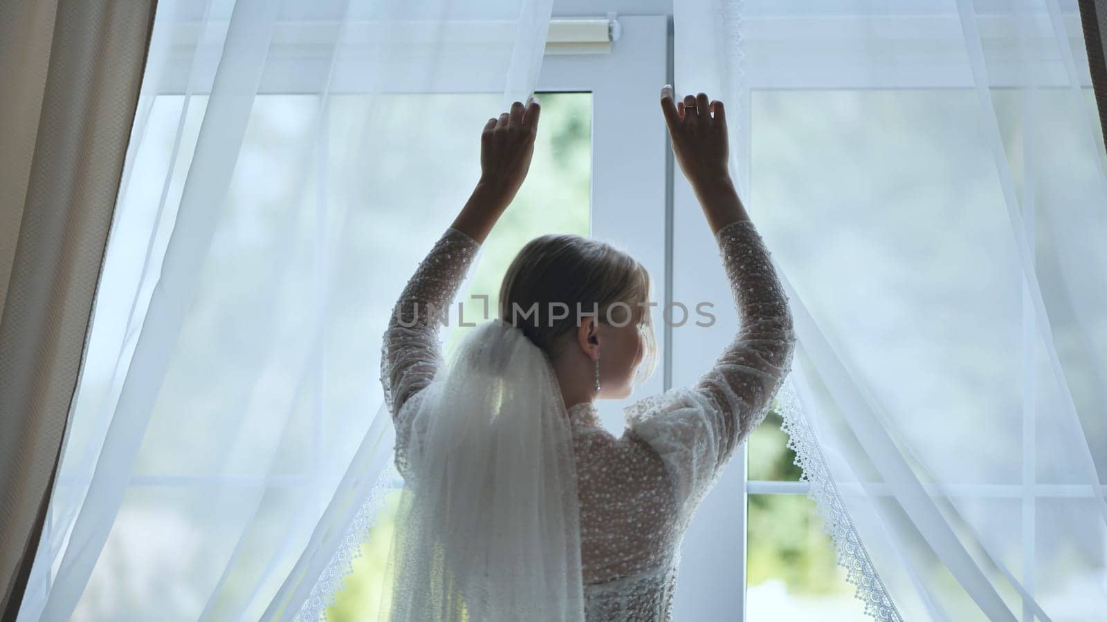 Bride, seen from behind, opens the curtains on her wedding day, revealing a beautiful green garden outside