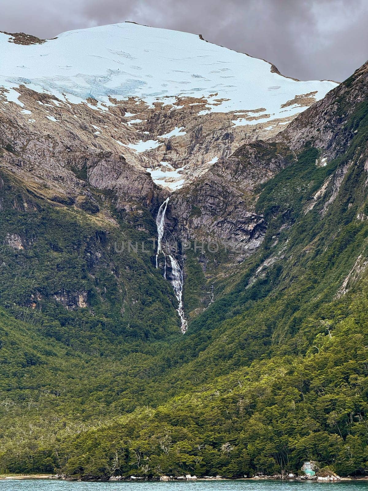 A narrow waterfall cascades from a glacier high in the snowy mountains, cutting through a forested valley and flowing toward a turquoise fjord below.