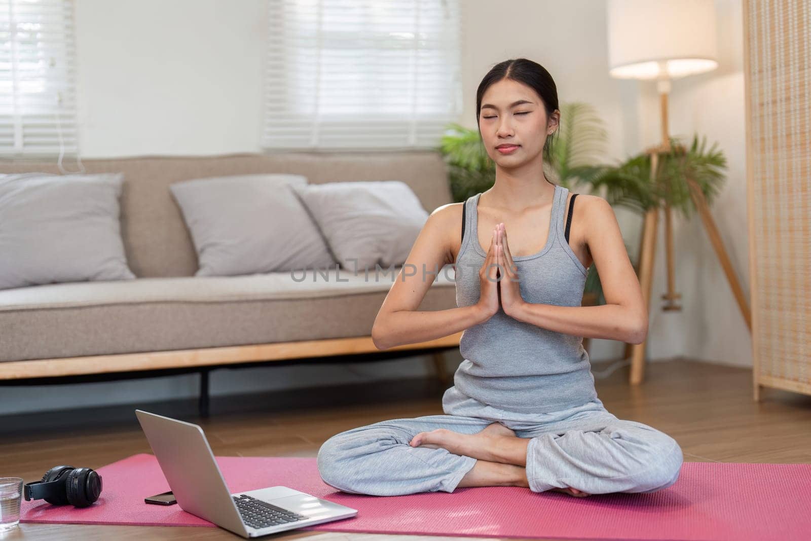 In a peaceful setting, a woman sits cross-legged on a yoga mat, practicing mindfulness and meditation. A laptop nearby provides guidance for her session.