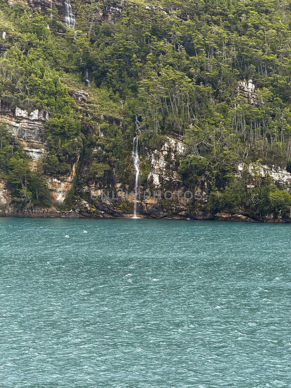 A delicate waterfall flows from a steep, green forested cliff into a vivid turquoise fjord, surrounded by rugged rock and dense vegetation.