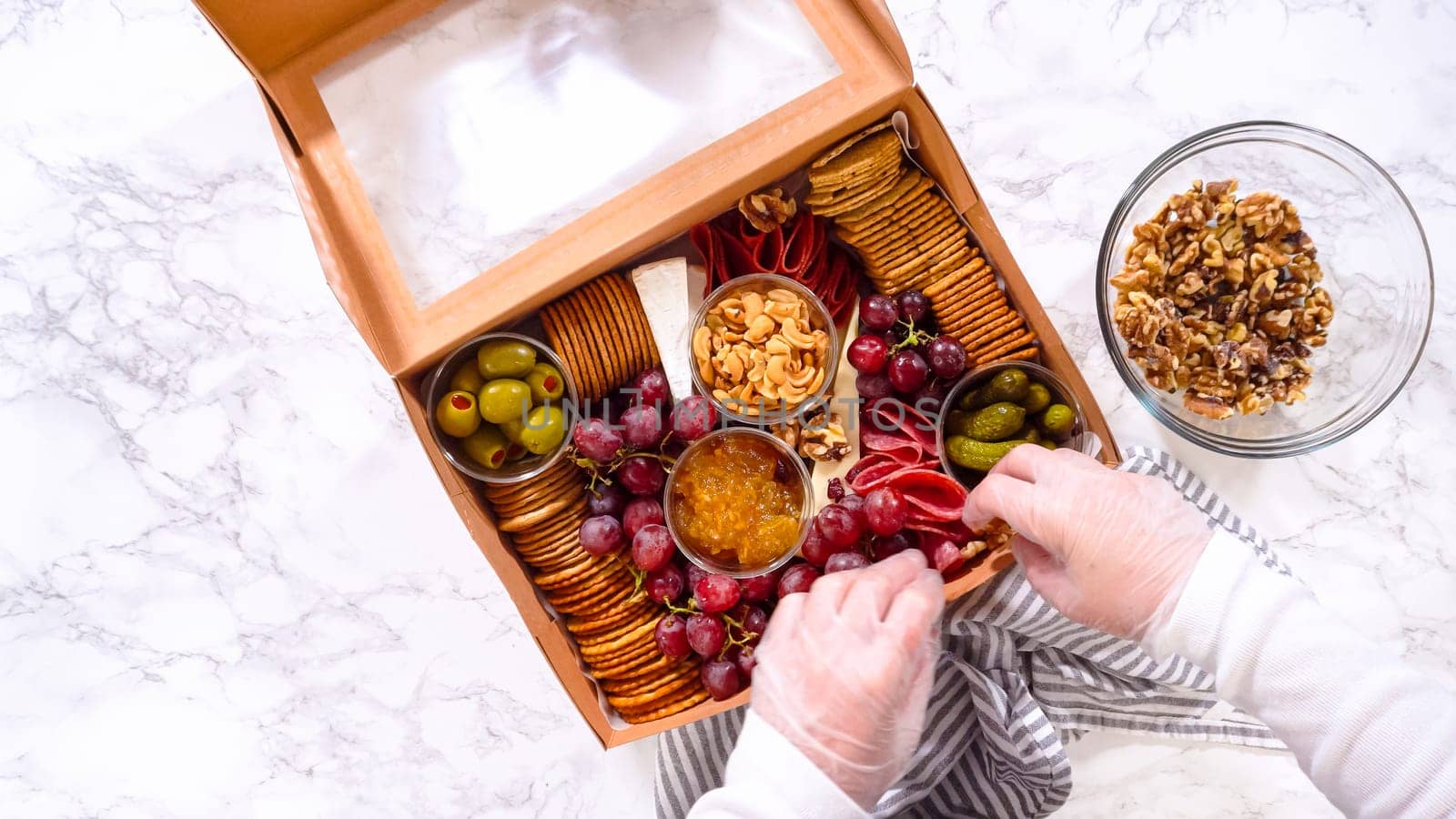 Flat lay. Hands are meticulously adding fresh red grapes to a bowl, complementing a beautifully arranged charcuterie box brimming with a variety of cheeses, olives, and cured meats, set against a sleek marble surface.
