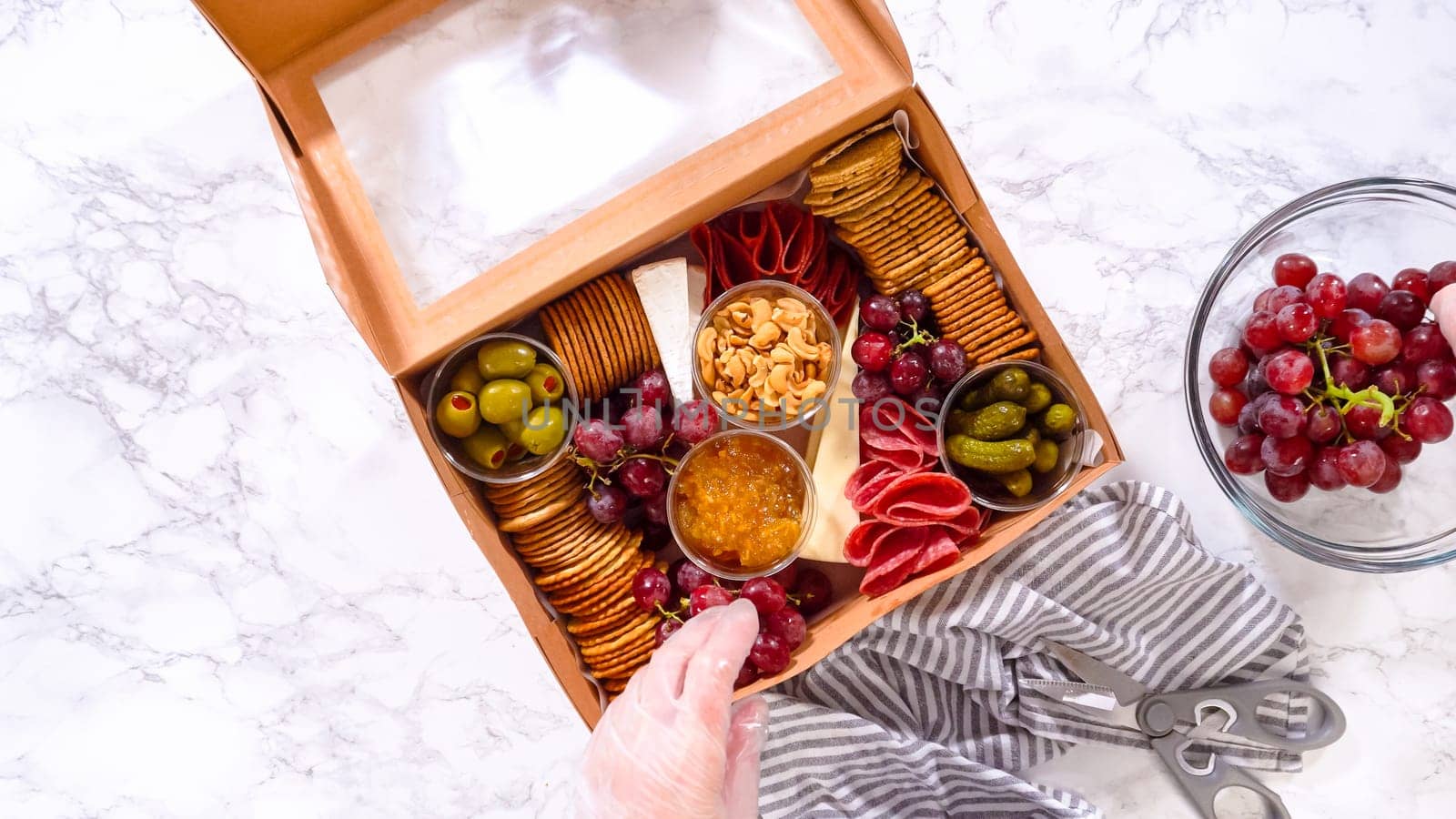 Flat lay. Hands are meticulously adding fresh red grapes to a bowl, complementing a beautifully arranged charcuterie box brimming with a variety of cheeses, olives, and cured meats, set against a sleek marble surface.