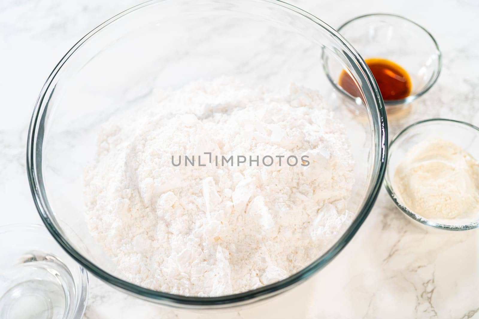 A glass bowl containing powdered sugar sits on a marble countertop, surrounded by small bowls of vanilla extract and other ingredients for preparing Simple White Royal Icing.